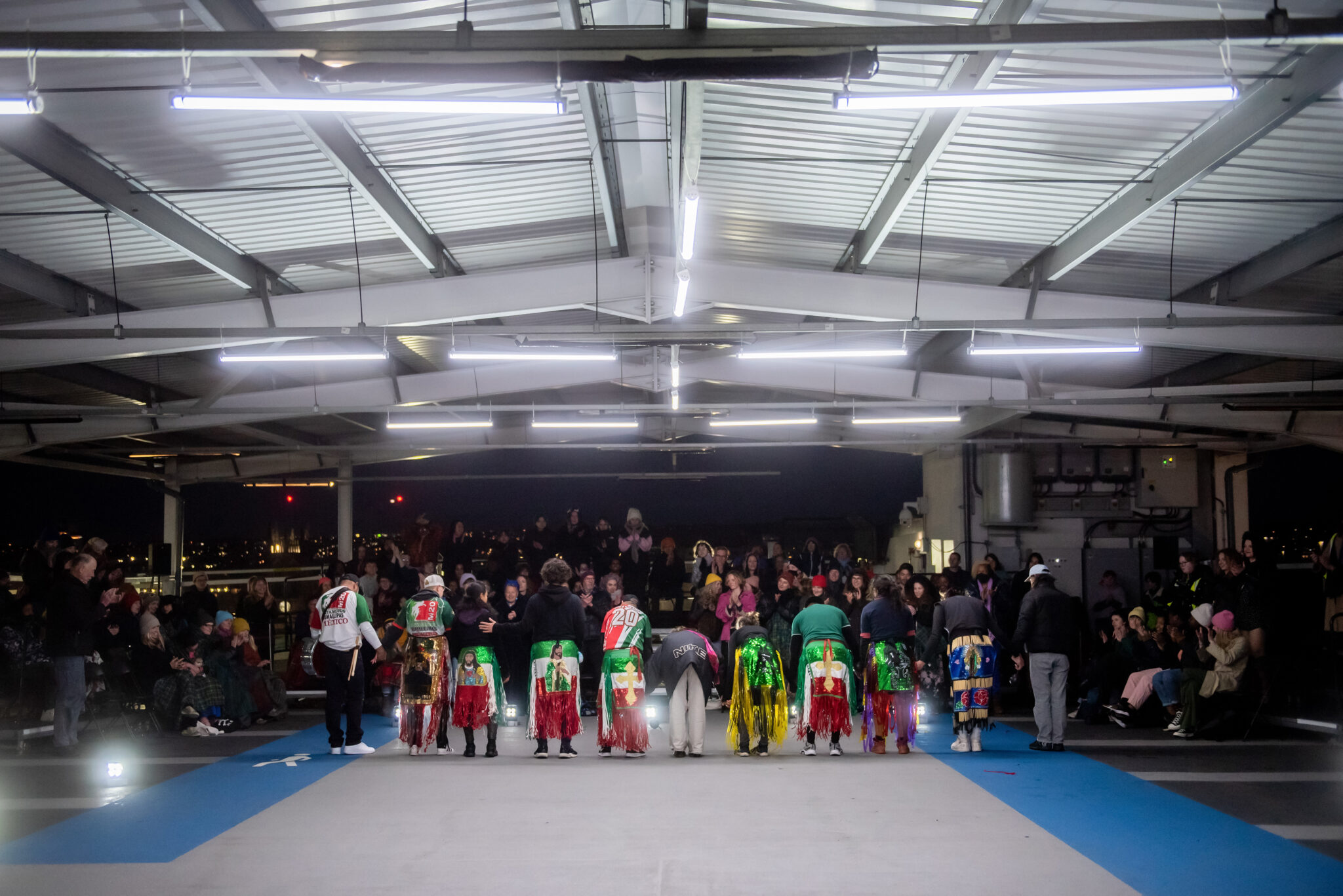 A group of performers in colourful, glittery costume stand with their backs to the camera. They are bowing and facing the audience who are clapping. They are standing on a grey concrete floor of a car park, with bright strip lights above them.