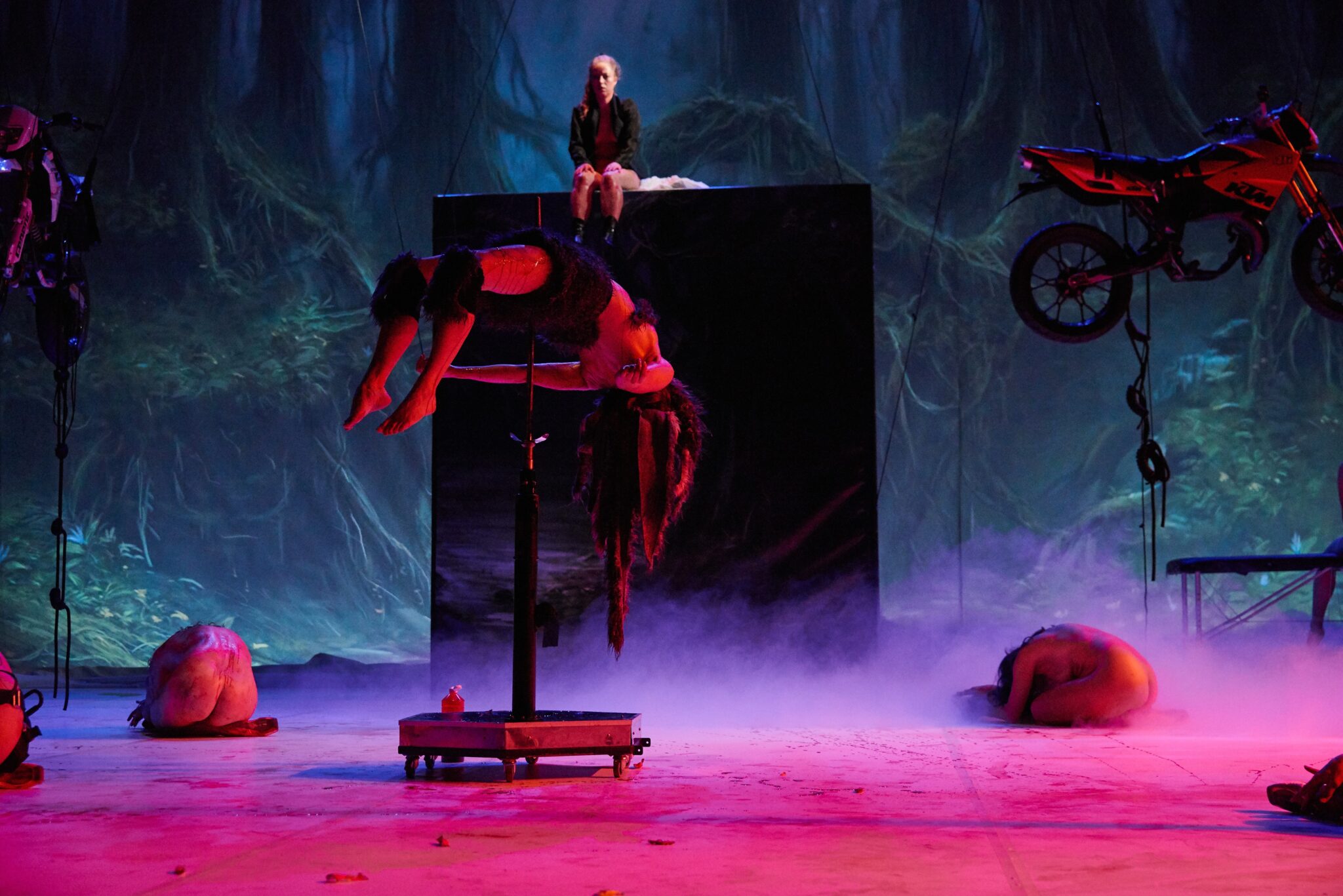 Performer lies draped across a podium in the centre of the image. They are wearing knee pads and something around their middle but nothing else. There are two naked women curled into balls on the floor facing away from the camera and one woman sitting towards the back of the image on a very large black box that takes up half of the image's background. There is a motorbike suspended on the right hand side. Lighting in this image is mostly red with a projection on the backdrop of a twisted dark forest, with tree roots entwined.