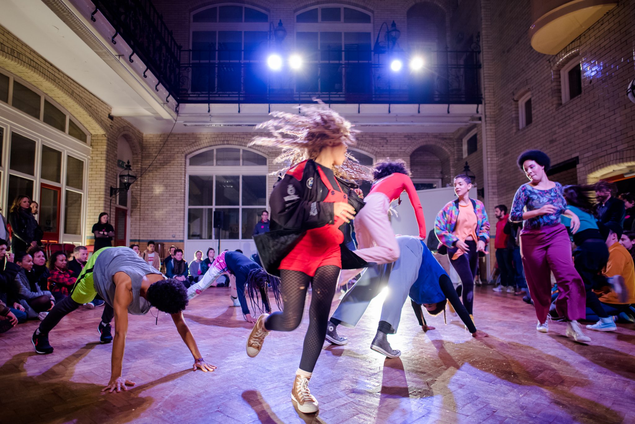 A group of performers dancing vigorously as part of a performance of When in Breaks it Burns. There is a woman in the foreground with red dungarees on who is mid-spin. The audience are watching from the floor around the space.