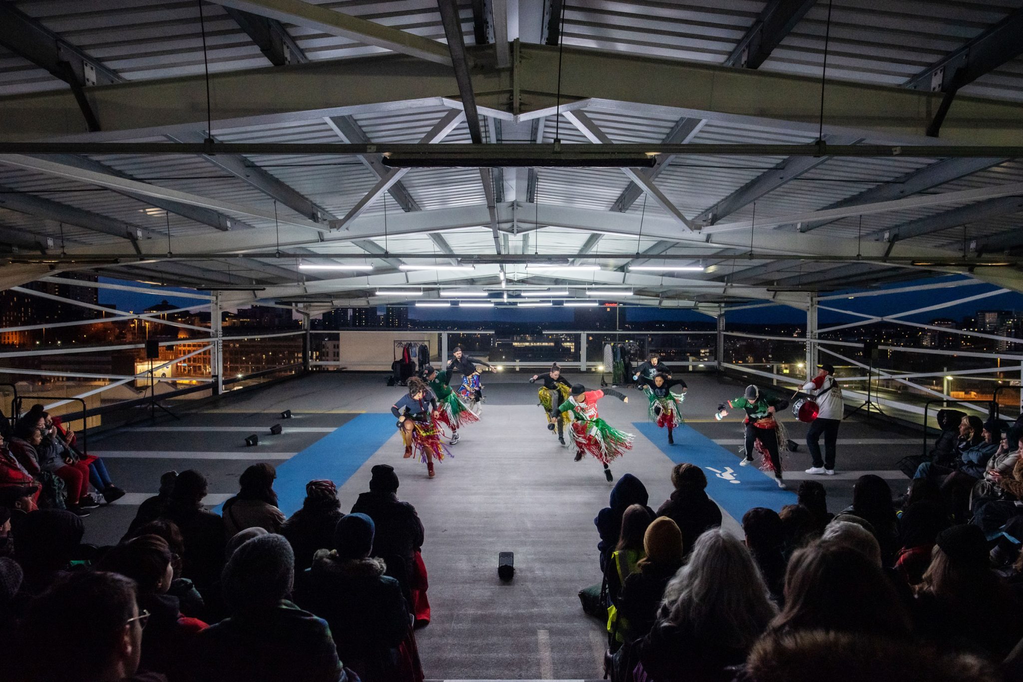 A performance unfolds on a car park with the sun setting on the Leeds city skyline